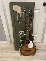Full view of the vintage Sears Silvertone acoustic guitar standing in front of the original box with visible branding and an attached shipping label.