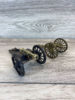 Two miniature metal cannons displayed side-by-side on a wood-textured surface. One has shiny brass barrel with black wheels, the other has bronze finish with bronze wheels.