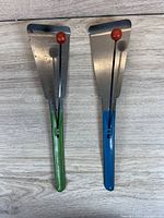 Front view of two vintage metal noisemakers with colored handles on a wood surface.