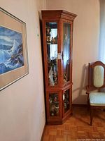 Wood corner cabinet in room corner with glass doors revealing shelves with collectibles inside. Light visible inside top section.
