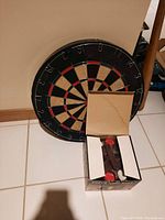 Photo of dartboard with a box containing darts in front, showing the dartboard face and boxed darts.