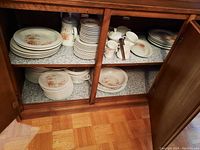 Wide view of wooden cabinet shelves with stacked dishes, cups, and pots marked with floral patterns showing the full dish set.