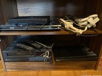 Four electronic media players on wood shelving, including Samsung Blu-ray, Toshiba DVD, Scott VCR, Sony CD/DVD, alongside remote controls and power bars.