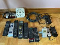 Various black remote controls, two older model cell phones with charging cables, and coiled audio/video cables on hardwood floor