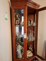 Front view of corner cabinet with three shelves filled with various figurines, glassware, and ceramic dishes.