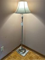 Full view of the floor lamp lit up, showing fabric shade and metal base stand against cornered wall.