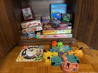 Front view of collection of boxed board games and tabletop games arranged on a wooden shelf and parquet floor, showing multiple game titles and colorful game components including four plastic hippos for Hungry Hungry Hippos.