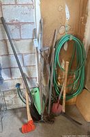 Full view of the lot showing coiled green garden hose on the wall and various hand tools leaning against the wall, including shovel, axe, rakes, pitchfork, spade, and orange knee pad on floor.