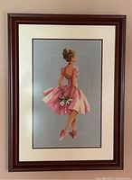 Full view of framed needlepoint artwork featuring ballerina in pink dress with dark wooden frame and cream matting.