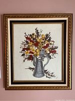 Framed needlework artwork showing full view of the needlework and frame with floral bouquet in pitcher.