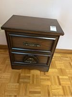 Front view of wooden nightstand showing two drawers with ornate metal handles and wood grain finish.