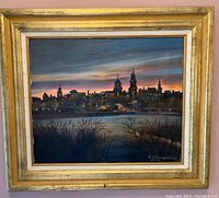 Full view of the signed and framed painting showing a city skyline at dusk with water and trees in foreground, signed by artist at bottom right.