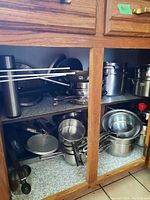 Kitchen cabinet with multiple stainless steel pots, pans, and strainers stacked and stored on shelves.