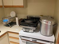 Photo of 4L aluminum stock pot with lid, stacked roasting pans, cast iron skillet, drying rack with plate, blue brush, and paper towel holder on kitchen counter and stove.
