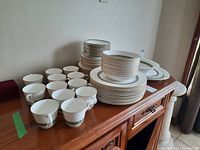 Full overview of many stacked plates, bowls, and cups grouped on a wooden cabinet showing the quantity and style of the dinnerware set