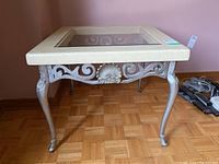 Front view of the square side table showing cream frame, glass top, and ornate metal designs on legs and side supports.