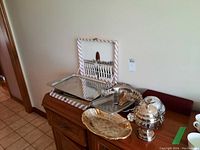 Overview photo showing various silver plate and stainless steel serving items on a wooden sideboard: napkin holder, serving trays, and bowls.