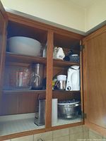 Photo of kitchen cabinet with Pyrex measuring cups, white electric kettle, mixer, grinder, milk pitcher, metal colander, mixing bowl, and glass bowl.