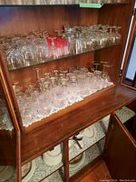 Overview of two shelves holding various stemware and glassware, including whiskey glasses, wine glasses, and shot glasses on a wooden cabinet shelf with lace liner.