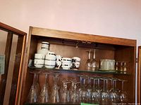 Wide view of one glass cabinet shelf showing stemware, coffee cups, and glasses stacked inside.