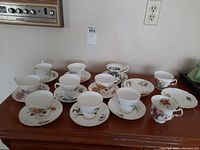 Wide view showing 11 teacups and 12 saucers arranged on a wooden table with various floral patterns and gold trim.