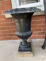 Front view of the black metal urn showing decorative oval pattern on the flared rim, ribbed neck and square base with stone slab underneath.