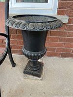 Full side view of black metal urn showing ornamental rim and square pedestal base on concrete ground.