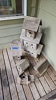 Full view of six weathered wooden birdhouses varying in size and shape, placed outdoors on a wooden surface against siding