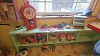 Full view of window shelf with vintage toys including pink toy clock, blue wooden toy piano, toy fire truck, doll, and other assorted toys