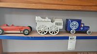 Red wind-up race car, white train, and blue Mar Toys truck displayed on shelf with auction tag