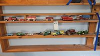 Full shelf with various model trucks and farming vehicles displayed horizontally on three wooden shelves, showing variety and condition.