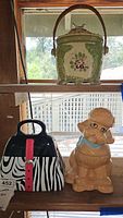 Shelf display of three ceramic cookie jars: top shelf lidded floral and bird jar, bottom shelf zebra-striped purse jar and tan poodle jar