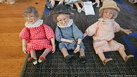 Wide shot showing all three elderly lady dolls arranged seated on a black mat, highlighting their facial features and dresses.