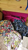 Three fabric bags on wooden surface: pink carry bag, blue paisley duffel, and beige-purple toiletry bag.
