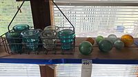 Blue and clear glass insulators arranged in a metal wire basket and blown glass globes in green, amber, and clear colors on a wooden surface by a window.
