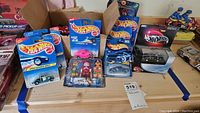 Photo of multiple Hot Wheels cars in packaging on wooden table, showing blue Volkswagen Beetle, pink car, police car and others in packages, also boxed Imaginext firefighter and Playmobil figures at rear