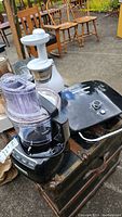 Photo showing a black KitchenAid food processor, a white Omega juicer, and a black George Foreman grill placed on a table outdoors.