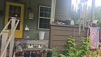Outdoor front porch area with yellow door and several weathered wooden birdhouses on steps and ledge, metal wind chimes hanging from post, various live plants in pots and trays on and around steps.