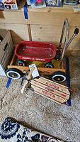 Photo of mini red Radio Flyer wagon and Classic Racer wooden sled on carpeted floor, showing the wagon's wheels, handle, and sled's wooden body with racer branding.