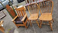 The lot photo shows the oak Mission style rocking chair with brown cushioned seat next to two oak spindle-back dining chairs on pavement outside.