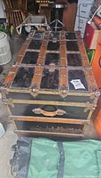 Front top view of vintage wooden trunk with black metal panels, leather handles and metal corner details showing wear and scratches.