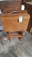 View of four wooden TV tray tables stacked on their stand. Medium brown wood with visible grain and cutout handles visible on the tray tops.