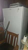 Front left angle view of white Frigidaire upright commercial freezer with door closed, showing handle and minor surface marks. Partially obstructed by wooden chair and miscellaneous items.