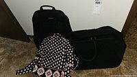 Photo showing three Vera Bradley travel bags: a black backpack, a large black duffle bag, and a patterned shoulder bag with red, white, and black motifs placed on the floor.