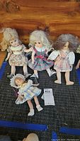 Four vintage dolls arranged on a dark cloth surface, showing full views of each doll including clothing and hairstyles. Dolls vary in size with one smaller doll wearing a bonnet and lace dress.