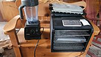 Vitamix 7500 blender and Magic Mill food dehydrator shown side by side on a wooden bench inside a garage setting.