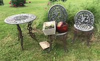 Overall view of cast iron outdoor bistro table and two chairs with open backs and ornate design, placed on grass with two planters on a chair