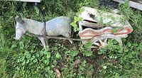 Concrete donkey with wagon sculpture resting on grassy area outdoors, tied together by metal rods to form a single piece. Shows presence of broken ear on donkey and chipped paint.