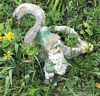 Concrete garden sculptures of a swan and a small gnome lying in grass and weeds, showing damage and weathering