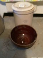 Full view of beige stoneware crock with lid and brown glazed bowl on floor.